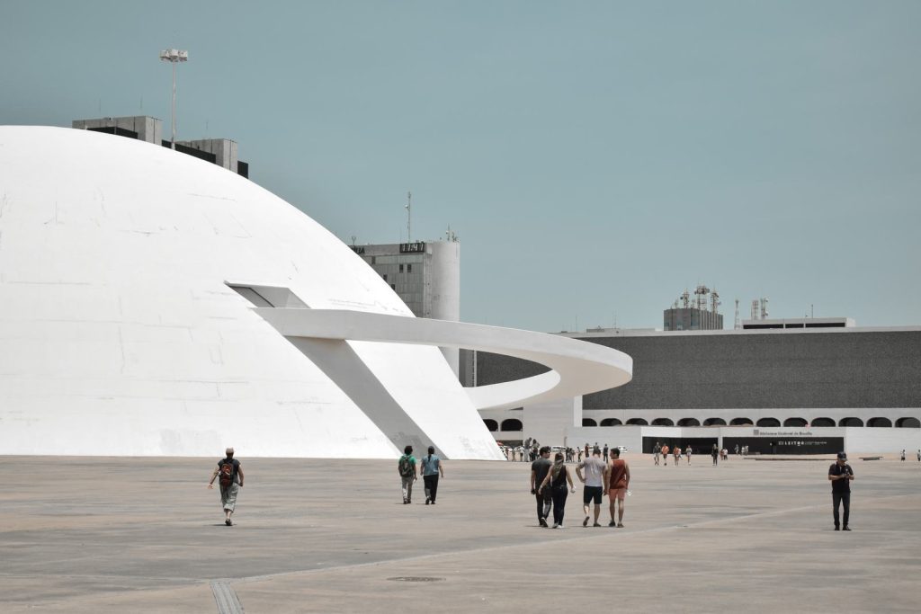 Cidade de Brasília, edíficio brando ao fundo com pessoas caminhando próximas à edificação. 